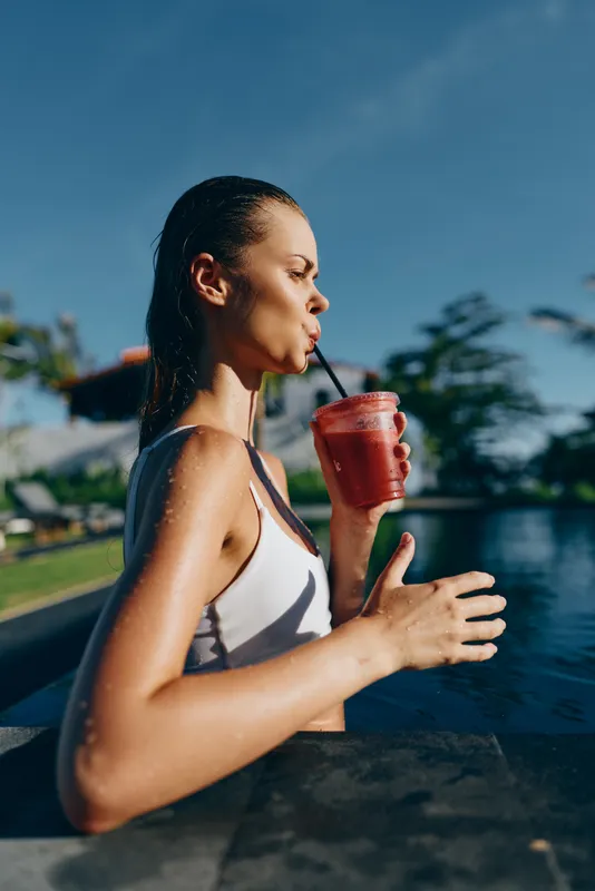 Young woman in white swimsuit drinks red smoothie while relaxing in outdoor pool on sunny day, enjoying healthy lifestyle and summer leisure time near water.