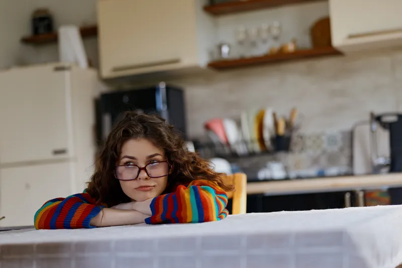Young woman with curly hair and glasses rests her chin on a kitchen counter, wearing a colorful striped sweater, looking thoughtful in a bright home setting. The scene feels candid.