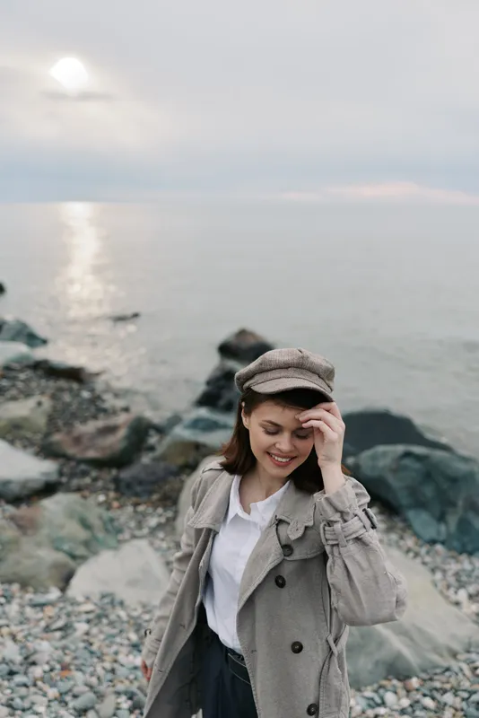Young woman in a coat and hat smiles while walking on a rocky beach during cloudy sunset. Relaxed mood with natural landscape and soft light near calm sea water.