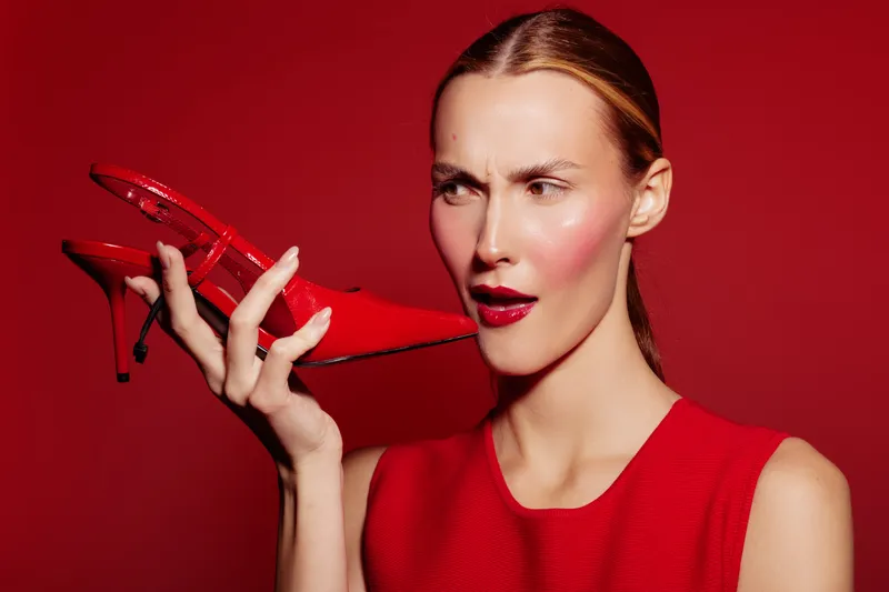 Young woman in red dress holds red high heel shoe near face with confused expression on red background. Concept of fashion, style, footwear, and emotions in studio lighting.