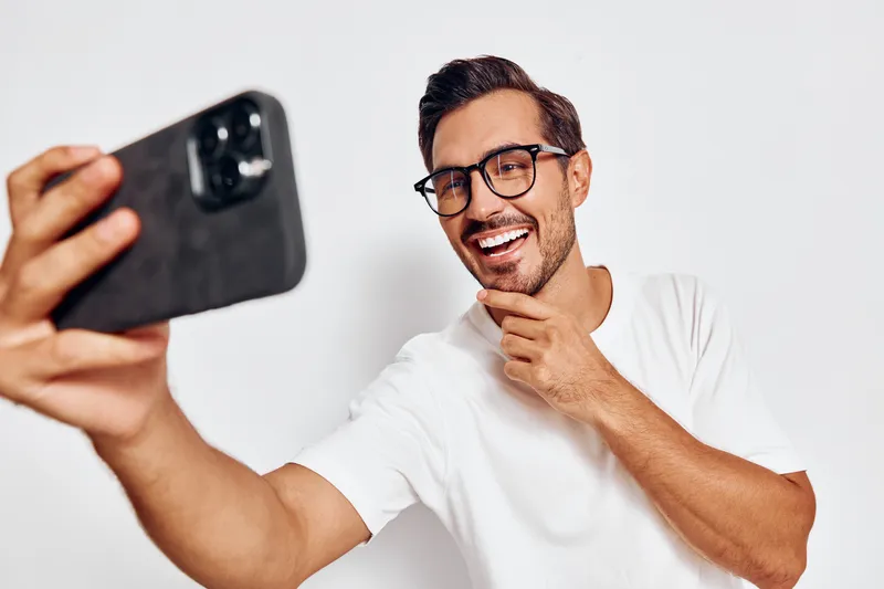 Smiling man taking a selfie with mobile phone, wearing glasses and white t-shirt, showing joyful expression against a plain white background. People lifestyle concept.