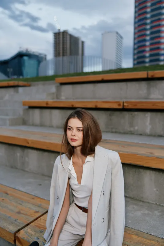 young woman in beige business suit sitting outdoors on wooden steps with urban background in autumn light, confident and thoughtful expression