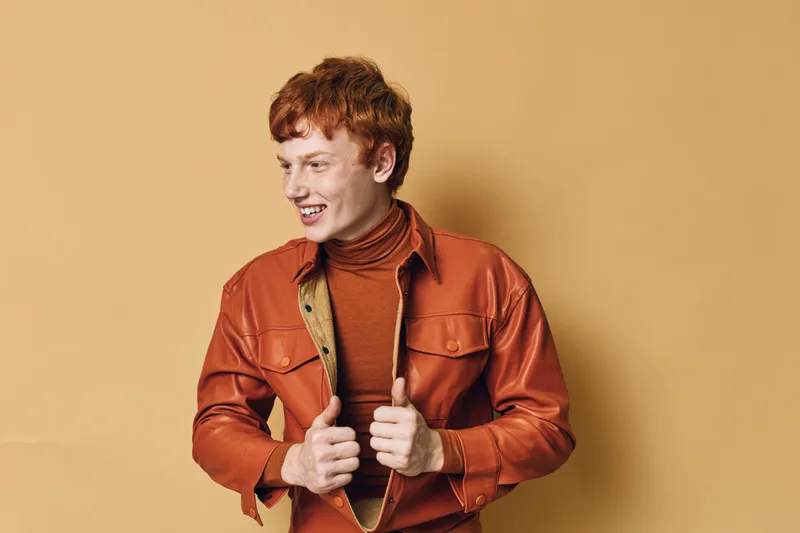 Young man with red hair wearing orange shirt and turtleneck smiling and looking to side on beige background in studio. Casual style with cheerful expression and fashion detail.