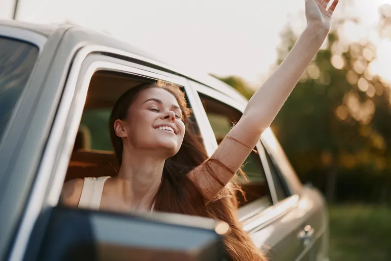 Young woman enjoying freedom with her arm out of the car window on a sunny day, evoking happiness and adventure in nature.