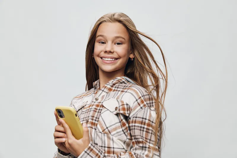 Smiling girl holding smartphone with yellow case wearing checked shirt with long hair in front of white background for technology and youth lifestyle concept.