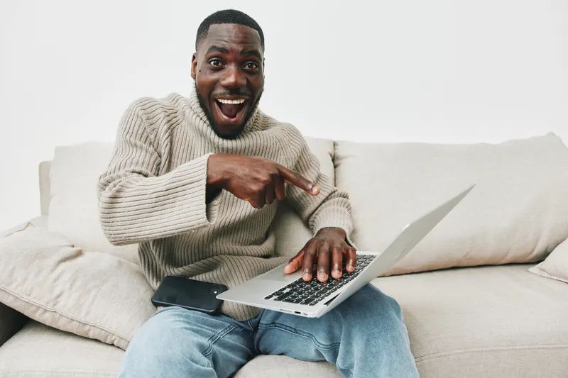 Happy young African man with laptop celebrating success at home, wearing a cozy sweater, conveying positive emotions, in a modern, bright living room