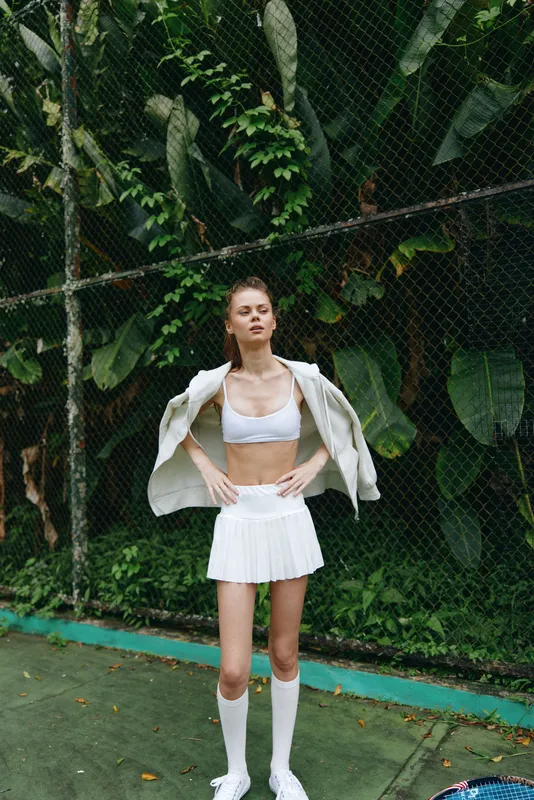 Young athletic woman in a stylish white outfit poses confidently on a tennis court surrounded by lush greenery, showcasing energy and elegance