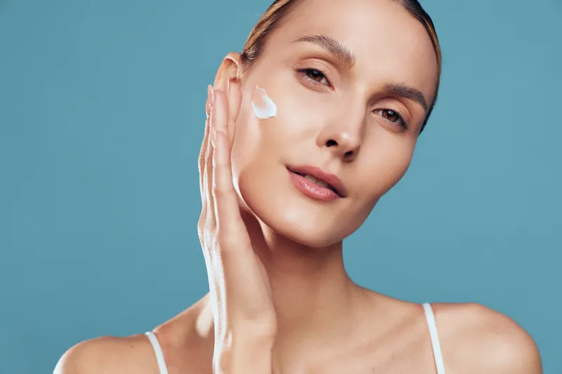 A young woman with clear skin applies white cream to her cheek against a blue background. Concept for skincare, beauty routine, hydration, and healthy skin care.