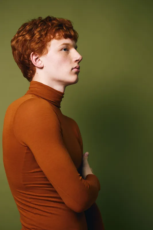 Portrait of a young man with red hair wearing a brown turtleneck against an olive background in profile with arms crossed looking forward thoughtfully.