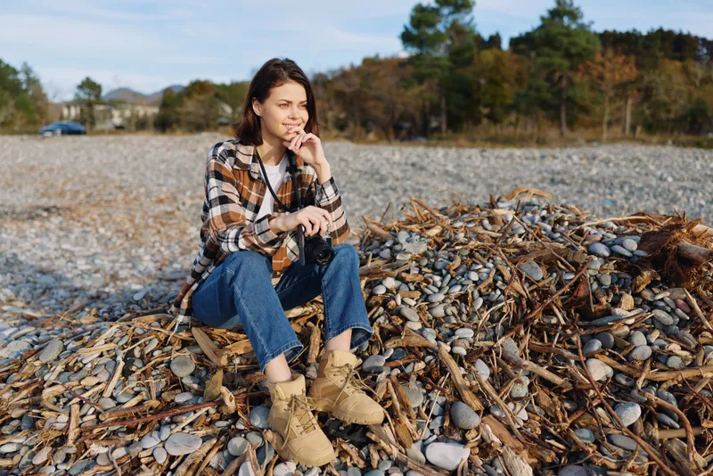Woman sits on a shell strewn beach, wearing a plaid shirt and rugged boots, enjoying a peaceful outdoor moment. Natural light highlights shells and a calm, reflective mood along the coast.