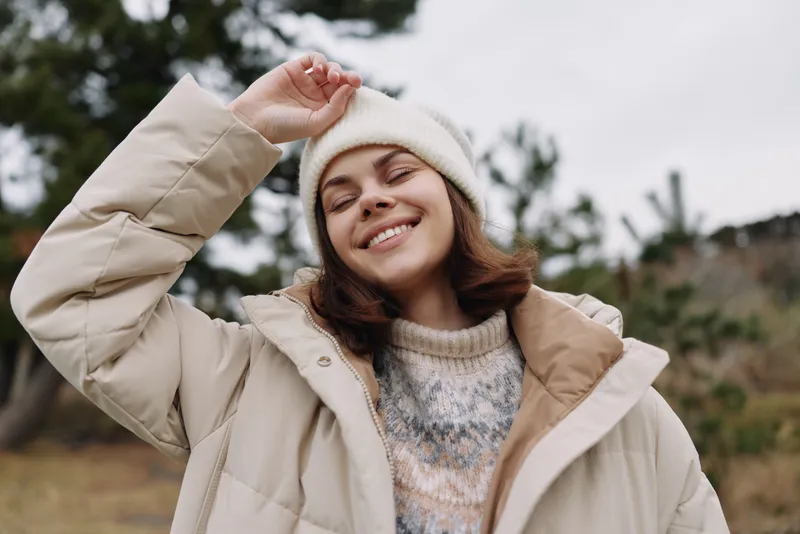 Outdoor portrait of a smiling woman in a cozy coat and knit sweater, wearing a beanie, enjoying fresh air in a cool setting with soft natural light and relaxed vibe.