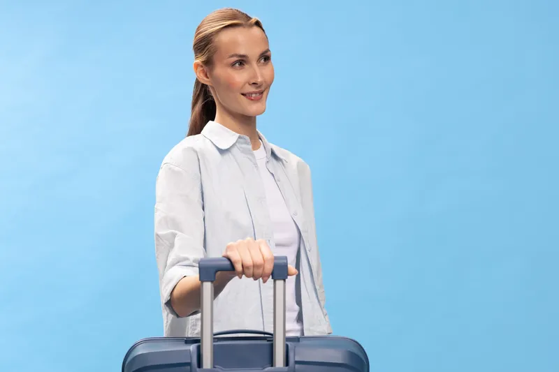 Young woman with suitcase smiling while standing against blue background. Travel concept with casual outfit and relaxed mood. Preparation for vacation or business trip.