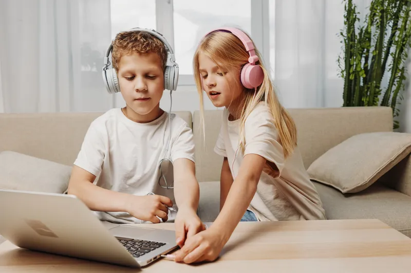 Two children with headphones use laptop at home, sitting on sofa. Concept of technology, learning, entertainment and childhood interaction.