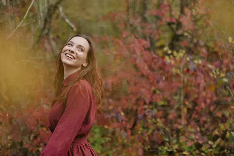 A genuine candid portrait of a smiling young woman in autumn foliage, capturing an authentic moment of joy and relaxed natural expression in warm, earthy tones and soft, flattering light.