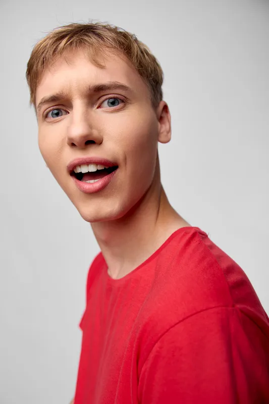 Close up of a young man in a red shirt with a surprised expression, captured in a bright studio setting, conveying genuine excitement and casual style suitable for lifestyle stock photography