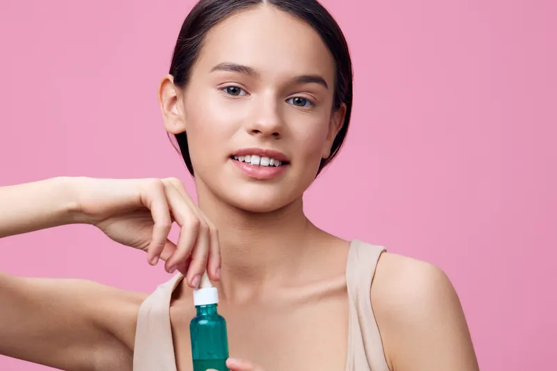 Young woman promoting skincare with a green serum on a pink background, showcasing natural beauty and a fresh, healthy look