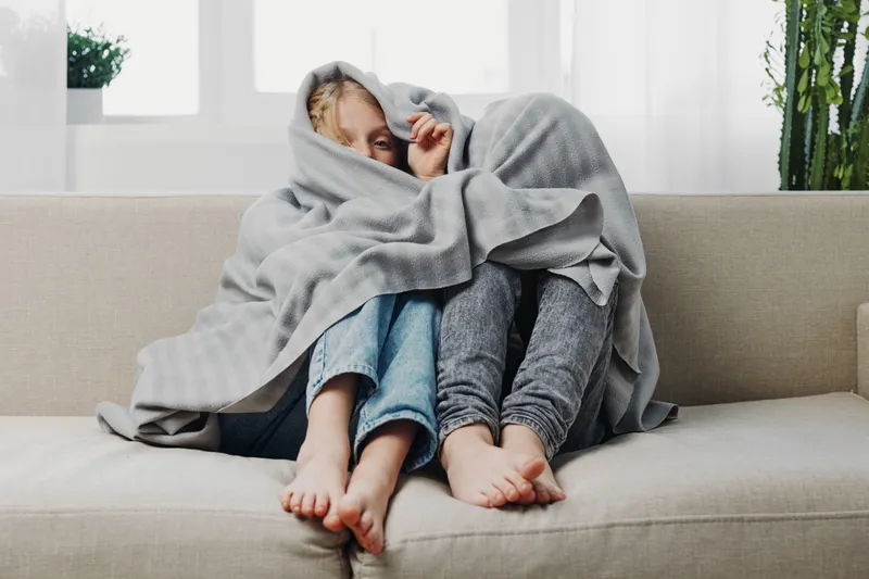 Two children wrapped in a grey blanket sit barefoot on a couch in a bright room. Cozy atmosphere with casual clothing and relaxed mood indoors.