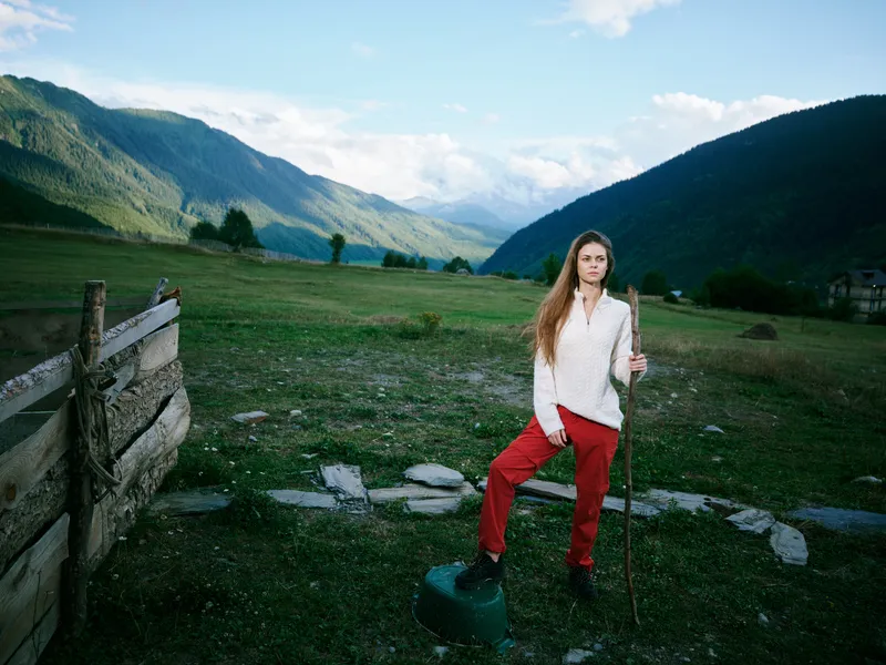 Valley landscape with distant mountains, a woman in white shirt and red pants stands beside a wooden fence in a rural field, outdoors, ready for hiking and exploration