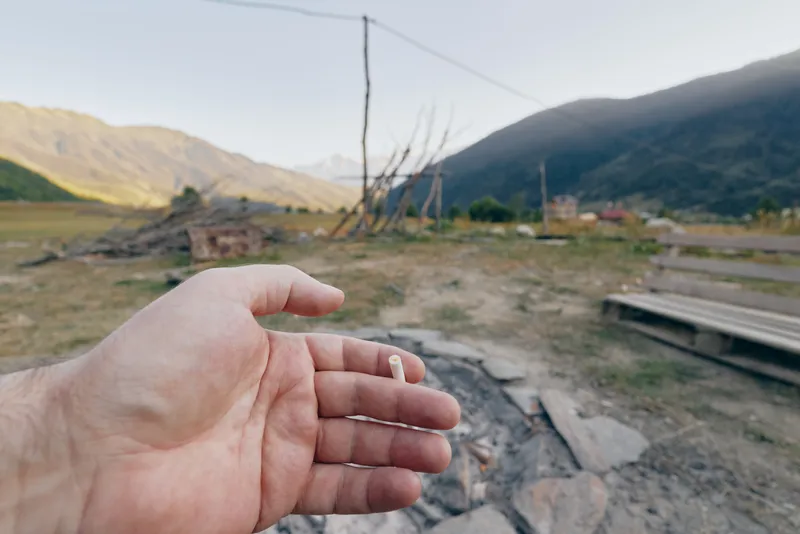 Hand cigarette outdoor mountains rural firepit smoke landscape valley field, closeup of a palm holding a spent cigarette butt near ash and ruined wood in a quiet mountain valley, daylight calm.