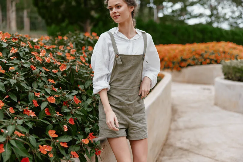 Young woman in stylish overalls smiling amidst vibrant flower garden, showcasing casual fashion and a joyful, sunny atmosphere in a botanical setting