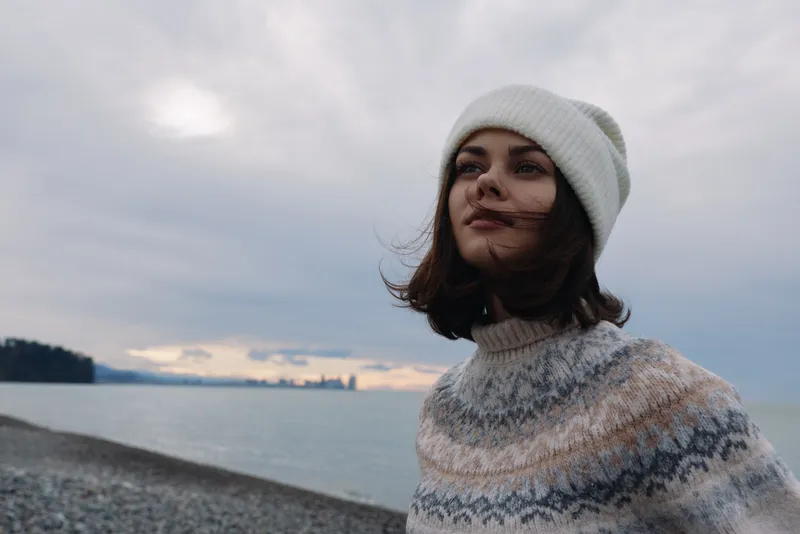 Portrait of a woman by the sea wearing a knitted sweater and white beanie, wind in her hair, calm coastline, moody clouds and soft light.