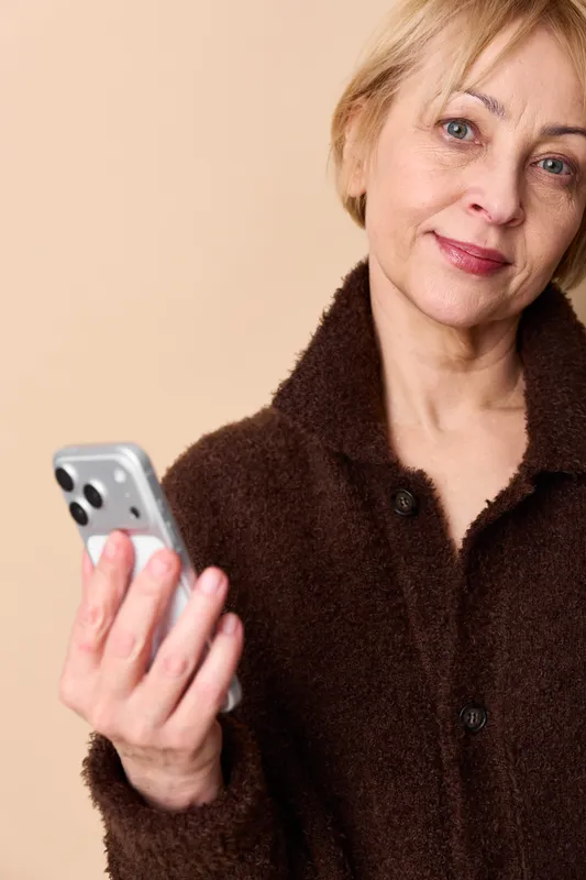 Studio portrait of a mature woman holding a smartphone, beige background, warm light indoors, brown coat, calm smile, relaxed hand, communication and technology