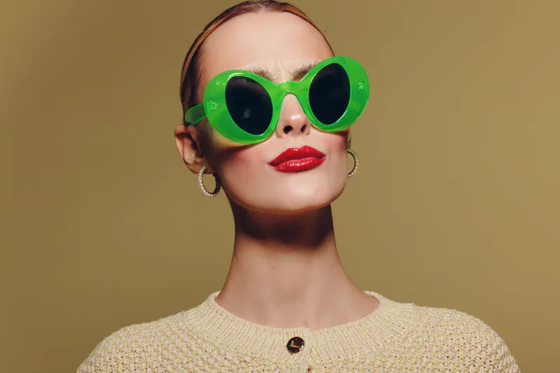Stylish woman wearing oversized green sunglasses and red lipstick poses confidently against beige background. Fashion portrait with trendy accessories and bold makeup concept.