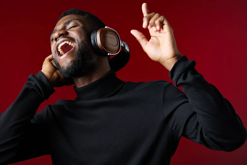 A joyful black man singing with headphones, wearing a black turtleneck, against a vibrant red background, expressing happiness and passion for music