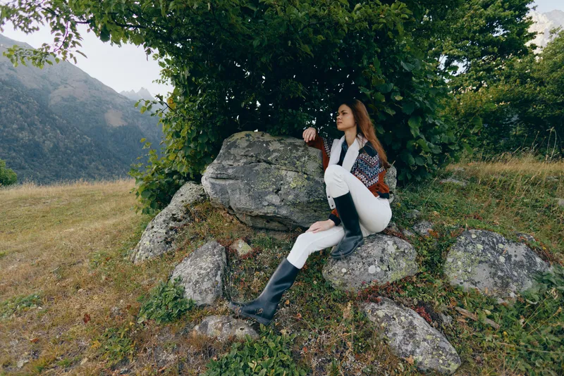 Woman sitting on rocks under a tree in a mountain meadow, wearing boots and sweater, enjoying nature and peaceful scenery with distant mountains and green grass.