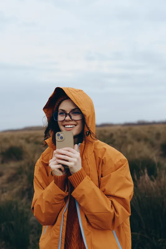 Woman lifestyle outdoors in autumn field, hooded orange coat smiling, holding smartphone for selfie, eyeglasses and relaxed expression, rural hills background, soft daylight, casual portrait