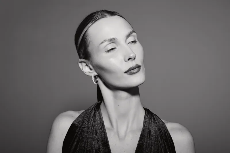 Elegant white woman with pale skin poses with eyes closed wearing shiny dress and earrings on dark background.