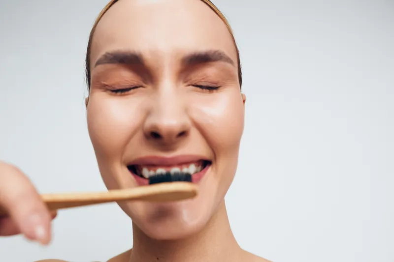 Smiling woman with closed eyes brushing teeth with bamboo toothbrush on white background. Concept of oral hygiene, dental care, natural lifestyle, and wellness. Soft natural lighting.