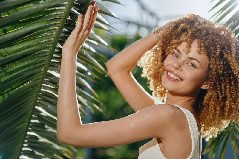 Smiling woman with curly hair enjoys sunlight behind tropical palm leaves in warm bright summer outdoors with natural green background.