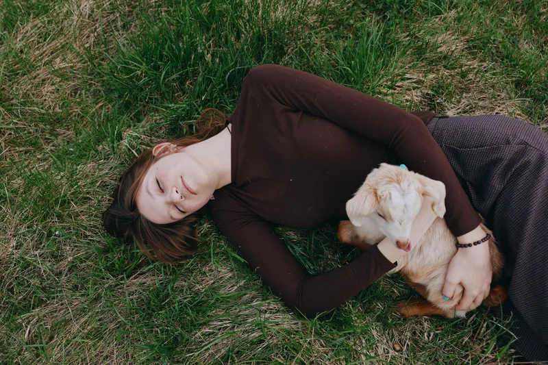 Cheerful woman outdoors lying on grass, cuddling a young goat kid, warm sunlight, relaxed posture, earthy tones, casual sweater, peaceful bond with animal, natural lifestyle moment