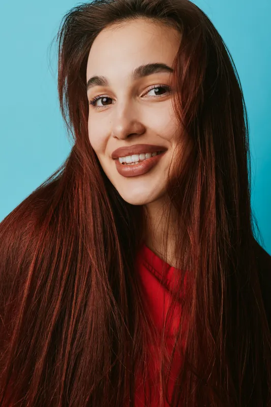 Woman with long brown hair and natural makeup smiling on blue background. Portrait of female with healthy skin and red clothing, happy expression and confident look in studio light.