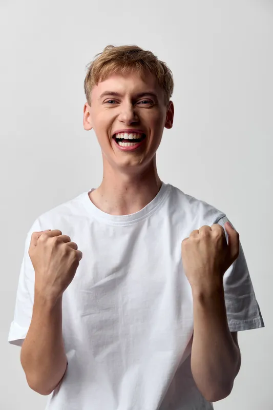Excited, joyful male in a white t shirt raises fists in celebration, bright smile and high energy conveying triumph, motivation, and confident enthusiasm for success.