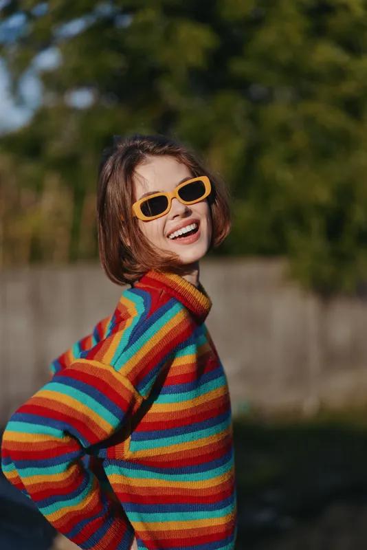 woman smiling striped sweater sunglasses outdoor young adult laughing in colorful rainbow knit jumper, short brown bob, yellow sunglasses, casual outfit in sunny backyard lifestyle portrait