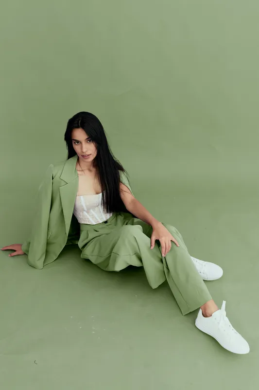 Stylish woman with long black hair wearing a green suit and white sneakers sitting on a matching green background, showcasing modern fashion and relaxed posture.