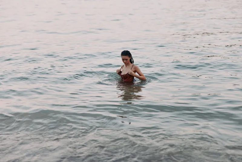 A woman stands waist-deep in calm ocean water, gazing downward, sunlit ripples surround her, capturing a serene moment of quiet reflection and connection with nature.