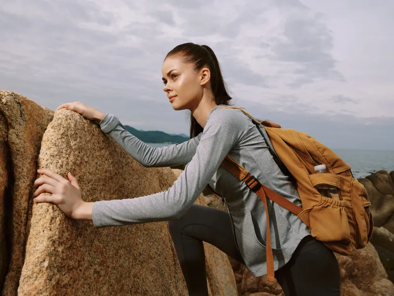 Young female hiker climbing rocks in nature, wearing gray athletic clothing and a brown backpack, showcasing determination and adventure against a cloudy backdrop
