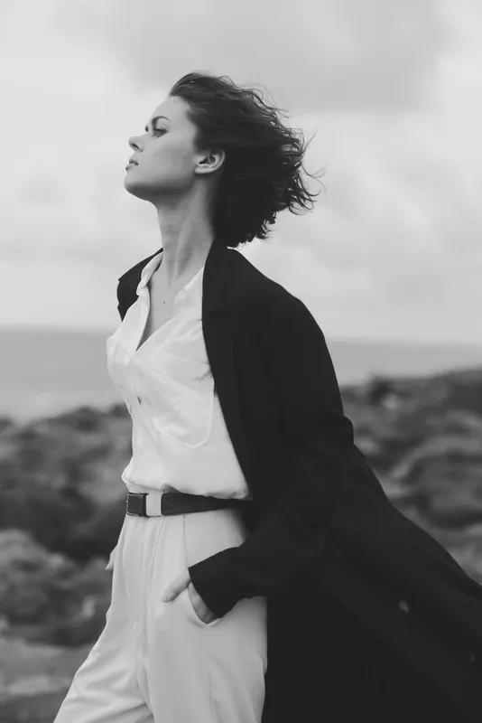 Woman walks along a windy coast in a flowing coat and white shirt, outdoor fashion mood with sea breeze, ocean backdrop, confident stride, dramatic atmosphere