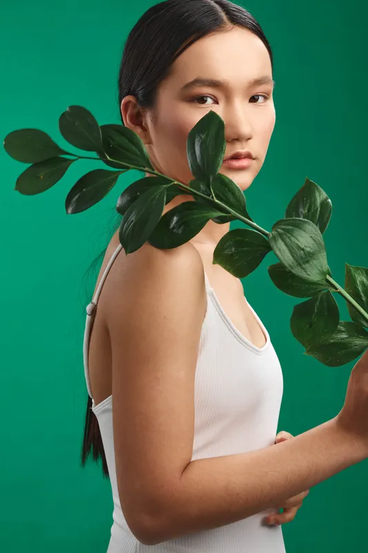 asian woman poses with a leafy branch against a green studio backdrop, creating a fresh natural aesthetic and serene mood. her calm expression enhances minimalist portrait vibes.