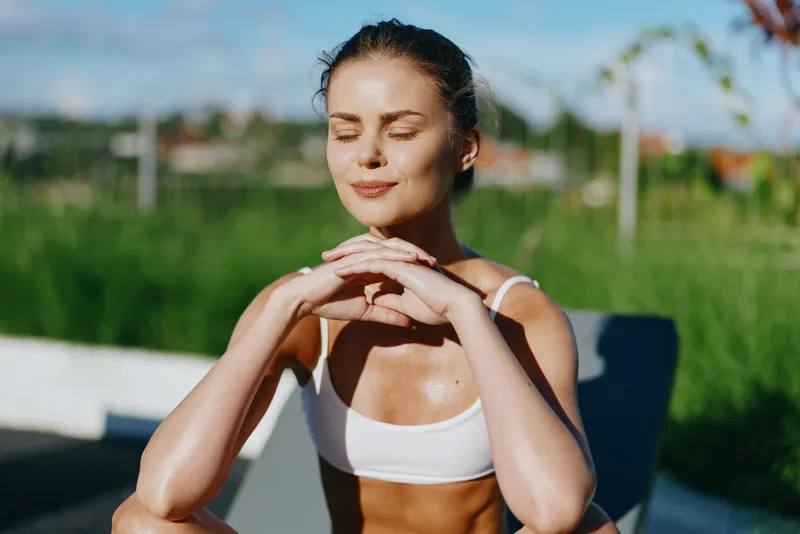 Relaxed woman outdoors, sunlit workout aftermath, eyes closed, gentle smile, hands clasped on knee, white sportswear, lush greenery in background, warm breeze, natural light, calm, candid lifestyle