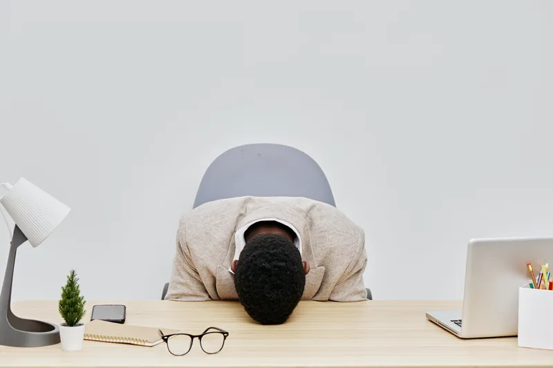 Frustrated young man with black hair leaning forward on desk, head down, distress, workplace stress, modern office environment, work-related pressure, feeling overwhelmed, tired mood.