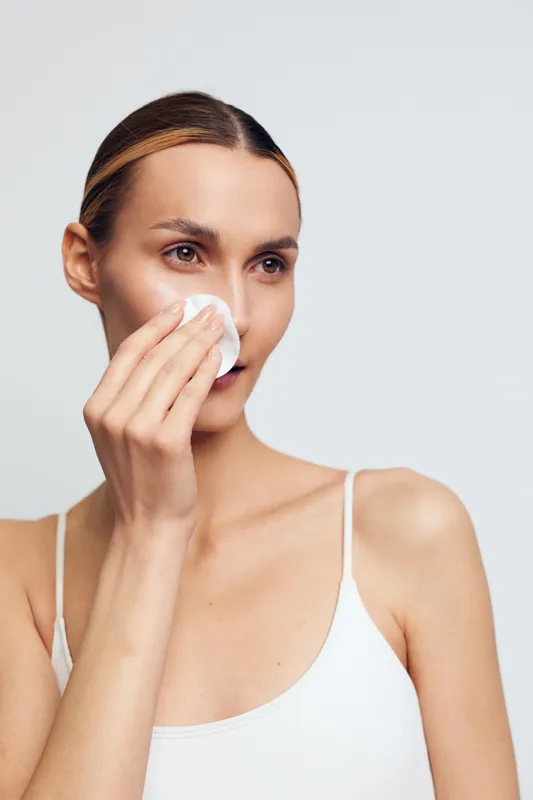 Young woman with light skin cleanses her face using a cotton pad, wearing a white tank top, captured in a calm skincare moment with soft lighting and a clean background.