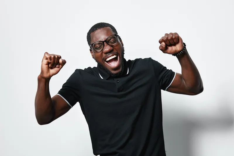 African American man with dark skin and glasses celebrating with raised fists and joyful expression wearing black polo shirt on white background. Happiness, success and excitement.