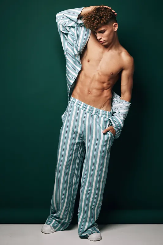 Man with muscular torso and curly hair wearing striped loose pants and shirt open standing against dark green background in artistic fashion posing with casual expression and one hand in pocket.
