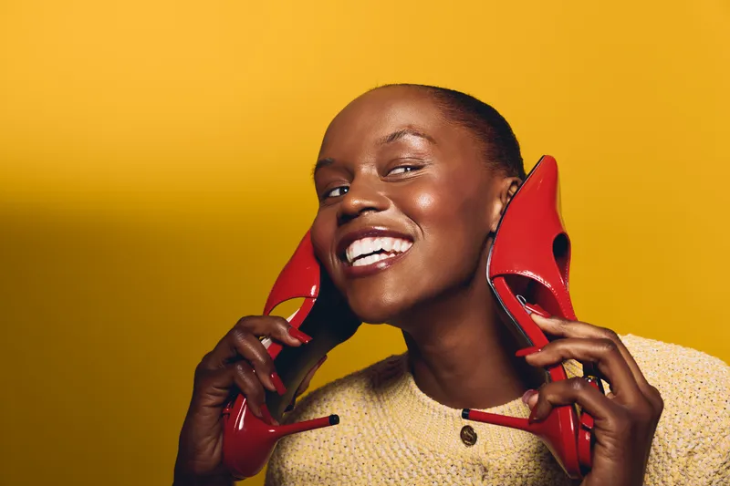 Smiling black woman with dark skin holds red high heels near face against yellow background. Concept for fashion, beauty, style, and joyful emotions in studio portrait.