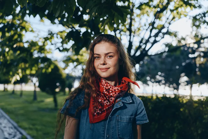 lifestyle woman in modern boho-western denim street style wearing a red bandana with natural sunlight and film color tones, outdoors in a green park setting