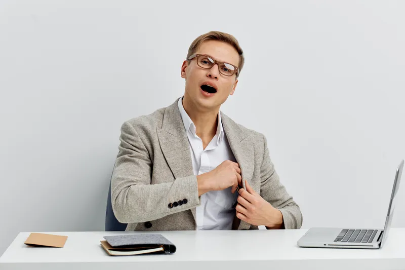 Confused young man in formal blazer and glasses sitting at a desk with laptop and notebooks, expressing surprise or realization. Studio shot with neutral background. People lifestyle concept, office w